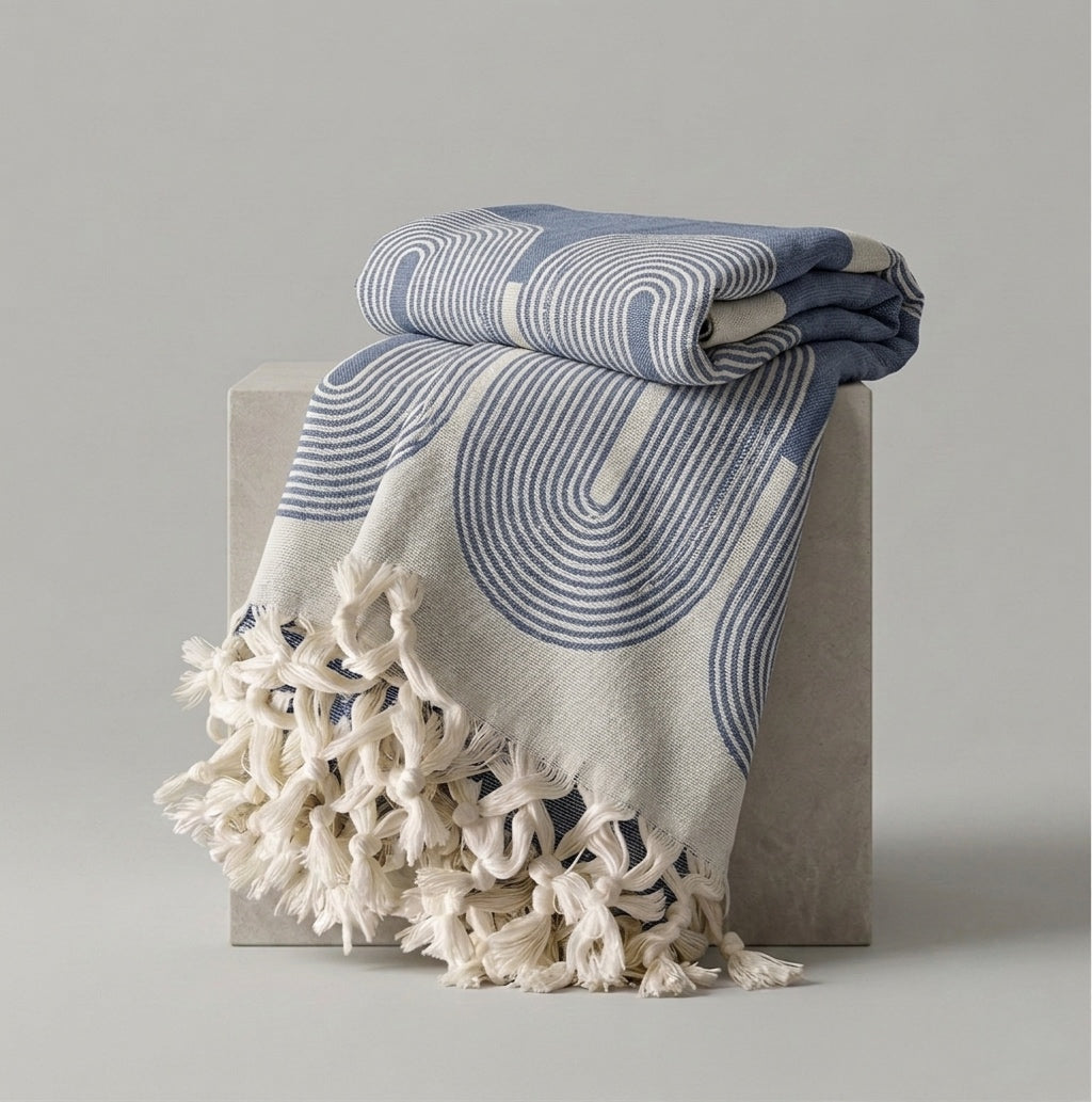 A folded and draped throw blanket with cream tassels, featuring a blue and cream wavy geometric pattern, displayed on a light gray block against a neutral gray background.