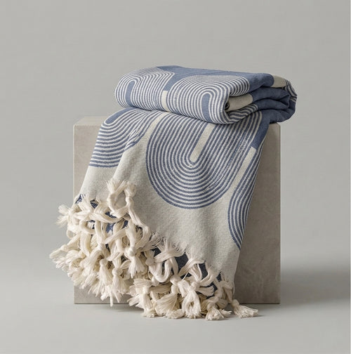 A folded and draped throw blanket with cream tassels, featuring a blue and cream wavy geometric pattern, displayed on a light gray block against a neutral gray background.